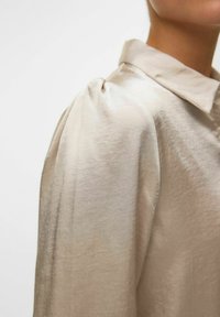 Beige bluse med krave, blød glans og samlede puffærmer. Glat tekstur og let materiale.