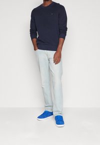 Homme portant un pull à manches longues bleu marine, un jean bleu clair et des baskets bleu vif, debout avec une main dans la poche devant un fond uni.