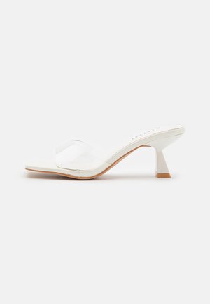 Witte open-toe mule sandaal met een doorzichtige band en een middelhoog uitlopend blokhakje op een effen achtergrond.