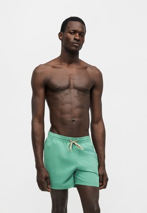 Άνδρας με σκούρο δέρμα και κοντά μαλλιά που στέκεται, φορώντας πράσινα μαγιό με κορδόνι, σε απλό ανοιχτό φόντο.