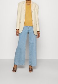 Femme portant un blazer beige sur un pull moutarde, un jean large bleu clair, et des talons noirs à brides, debout devant un fond blanc.