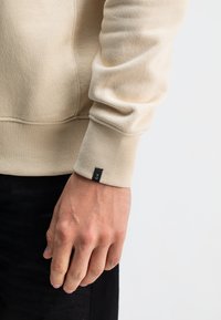 Beige sweatshirt met geribbelde manchetten, voorzien van een klein zwart label op de pols. Soepele textuur en een relaxte pasvorm, gecombineerd met donkere jeans.