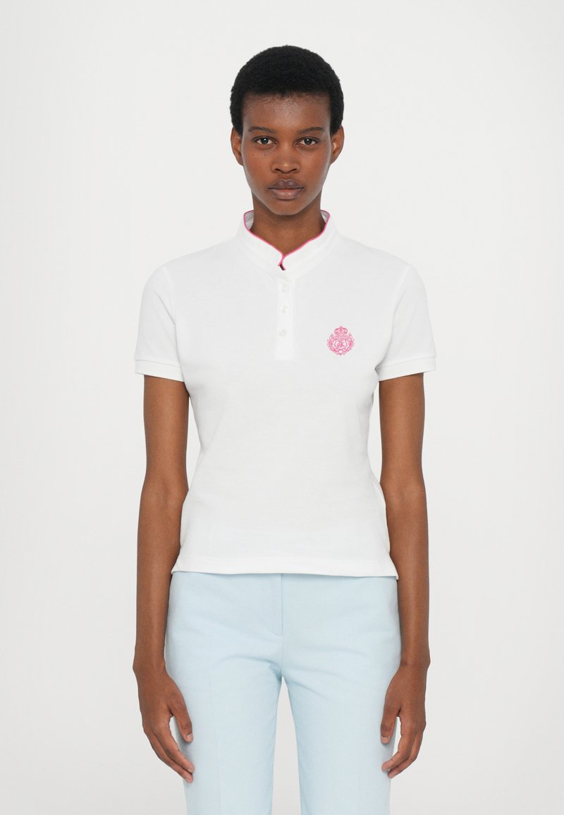 Hvitt bomullspolo med kort mandarin-krage, rosa detaljer og et lite brodert rosa våpenskjold på venstre bryst.