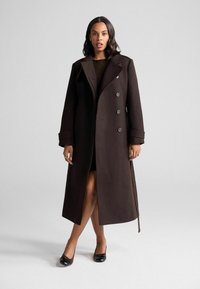 Lungo cappotto marrone scuro con colletto, bottoni doppi e maniche larghe. Texture liscia con una spacco laterale e un tailoring sottile.