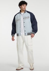 Lichtblauwe varsityjacket met marineblauwe mouwen, witte gestreepte manchetten en "Guess Jeans" geborduurd op de voorkant; gecombineerd met witte cargobroek.