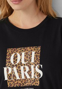 T-shirt noir avec un carré en imprimé léopard et le texte blanc « OUI PARIS ». Col rond avec une texture de tissu douce.
