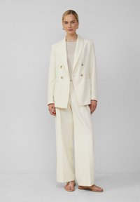 Dubbelzijdig witte blazer met gouden knopen, gecombineerd met brede witte broeken, aangevuld met een lichtbeige top en tan kleurige sandalen.