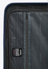 Interno nero di una valigia che presenta tasche in rete, scomparti con cerniera e un'etichetta di marca su uno sfondo grigio con motivo a righe.
