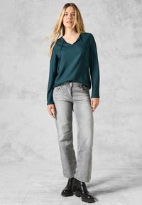 Maglia lunga verde acqua con scollo a V e coulisse. Jeans grigi dritti con una finitura scolorita. Stivali neri con suola spessa. Sfondo texturizzato.