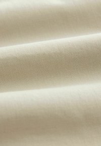 Gros plan sur un tissu beige lisse à texture tissée en diagonale, avec des plis doux créant un contraste de lumière et d'ombre.