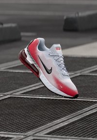 Sneaker Nike Air Max bianco e rosso con logo Swoosh nero, esposto in un angolo su griglie metalliche all'aperto.