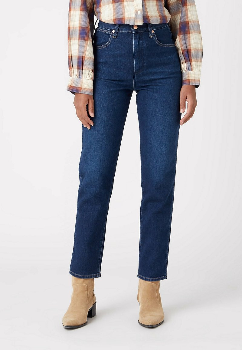 Jean bleu foncé taille haute à coupe droite, en denim avec des surpiqûres contrastantes, associé à une chemise à carreaux et des bottes beiges.