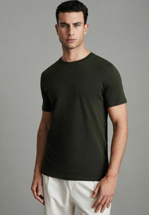 Olivgrünes, kurzärmliges Baumwoll-T-Shirt mit Rundhalsausschnitt; glatte Textur und taillierter Schnitt; getragen mit hellen Hosen.