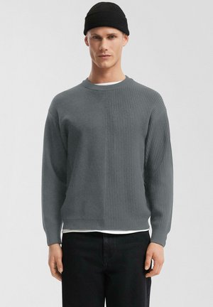 Mann trägt einen grauen, gerippten Pullover, weißes Unterhemd, schwarze Hose und eine schwarze Mütze, steht vor einem schlichten hellen Hintergrund.