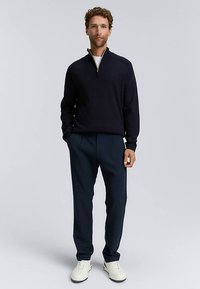 Mörkblå pullover med halvlång dragkedja, kombinerad med skräddarsydda mörkblå byxor och vita sneakers. Mjuk textur, enkel design och figurnära silhuett.