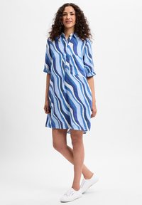 Blaues Leinenkleid mit wellenförmigen Streifen in Hell- und Dunkelblau, mit einem Knopfkragen und einer einzelnen Brusttasche. Getragen mit weißen Sneakers.
