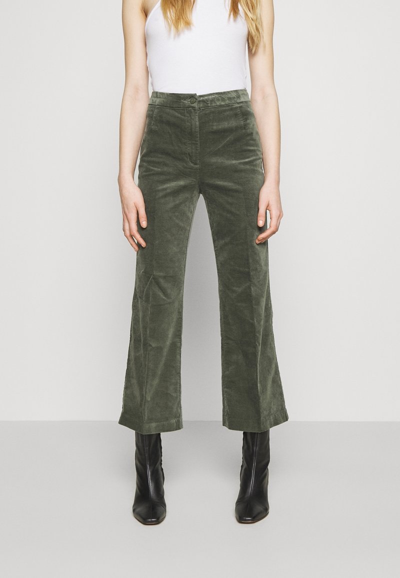 Femme portant un pantalon large en velours côtelé vert olive taille haute avec bouton et fermeture éclair à l'avant, bottines noires et haut blanc sans manches.