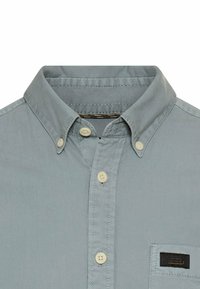 Helles blaugraues Hemd mit Kragen, ausgestattet mit cremefarbenen Knöpfen und einer Brusttasche mit einem kleinen Markenetikett. Glatte Textur.