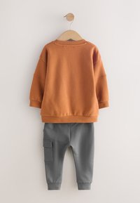 Sweat-shirt orange à épaules tombantes, associé à un pantalon de jogging gris avec une poche latérale. Les deux vêtements sont fabriqués dans un tissu doux et confortable.