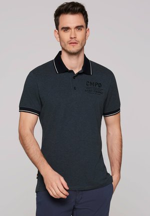 Mann trägt ein dunkelgraues Poloshirt mit schwarzem Kragen und Ärmelsaum, steht mit einer Hand in der Tasche vor einem schlichten Hintergrund.