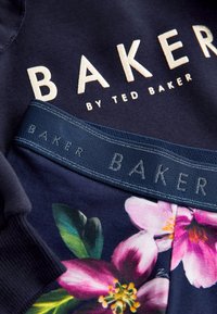 Tumesinine puuvillane dressipluus, millel on broderitud "BAKER" logo, koos lillemustriliste lühikesed püksidega, millel on sinine elastne vöökoht.