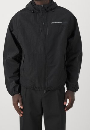 Homme portant une veste à capuche zippée noire EA7 Emporio Armani avec des poignets élastiques et des poches latérales, associée à un pantalon noir.