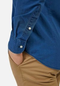 Camicia button-up blu scuro con una texture liscia, caratterizzata da polsini con bottoni e un orlo leggermente curvo, abbinata a pantaloni marrone chiaro.