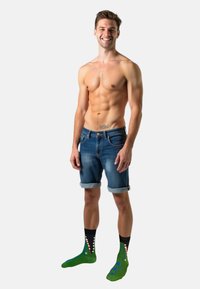 Mand iført oprullede denimshorts og farverige sokker med krokodille-design, der viser grønne, sorte og hvide farver.