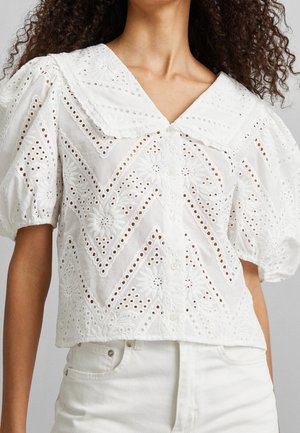 Hvid blondebluse med øjlemønster og pufærmer, blomstermotiver og geometriske udskæringer, stor spids krave, knappet front, båret sammen med hvide bukser.