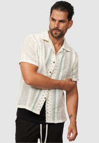Weißes kurzärmliges Hemd aus offen gestricktem Stoff mit grünen Streifen und Kragen. Verfügt über eine Knopfleiste und einen lässigen Schnitt. Schwarzes Unterhemd sichtbar.