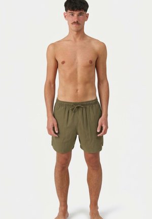 Młody mężczyzna z krótkimi brązowymi włosami i wąsami, stojący boso, ubrany w oliwkowo-zielone kąpielówki z troczkiem, na jednolitym białym tle.