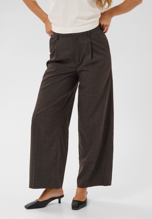 Femme portant un pantalon marron à carreaux à jambes larges et des chaussures noires basses à enfiler avec un haut blanc sans manches.