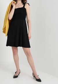 Černé šaty s nařaseným živůtkem, nastavitelné ramínka a rozšířená sukně. V kombinaci se žlutým svrchním oděvem a černými podpatky se mašličkami.