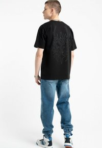 Czarny bawełniany t-shirt z dużym, skomplikowanym wzorem graficznym na plecach. Połączony z jasnoniebieskimi dżinsami i trampkami z niebieskimi i czarnymi akcentami.