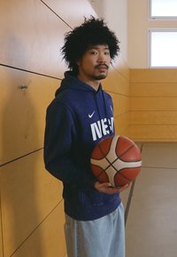 Mėlyna gobtuvas su balta "NIKE" užrašu, šviesiai mėlynos džinsai, laiko krepšinį. Fone matoma medinė sporto salės siena ir langai.