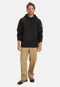 Felpa grigia scura con tasca frontale, cappuccio regolabile con cordini e un'etichetta in pelle marrone. Abbinata a pantaloni beige e sneaker scure.