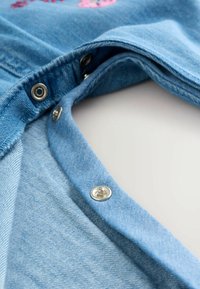 Világoskék farmeranyag ezüst szempánt gombokkal a nyaklánc mentén. Strukturált felülettel és látható varrási részletekkel rendelkezik.