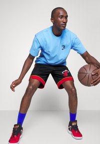 Svetlomodré tričko, čierne basketbalové šortky s červenými a žltými akcentmi a červené tenisky. Hráč drží basketbal, pripravený na dribling.