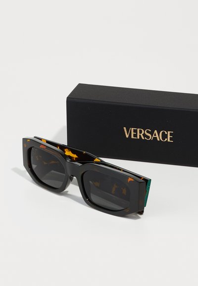 Black sunglasses with orange tortoiseshell accents and square frames, positioned next to a black Versace box with gold text.