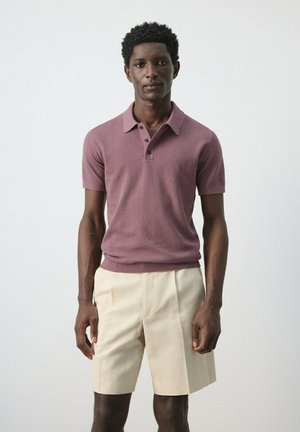 Hombre vistiendo una camisa polo de manga corta color malva y pantalones cortos beige hasta la rodilla, de pie sobre un fondo blanco liso.