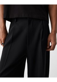 Černé kalhoty na míru vyrobené z hladké tkaniny, s ostrými záhyby, slim fit v pase a jemným leskem. Ruka s viditelným stříbrným prstenem.
