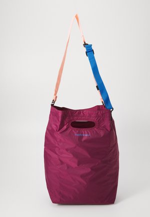 Grand sac fourre-tout bordeaux avec une découpe pour la poignée supérieure, une bandoulière rose et une sangle bleue réglable suspendue sur fond blanc.