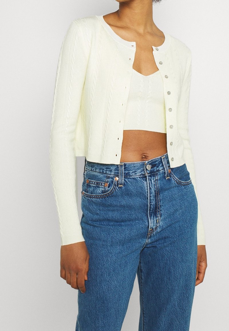 Femme portant un cardigan court blanc par-dessus un crop top en maille blanche avec un jean en denim bleu taille haute sur un fond clair.