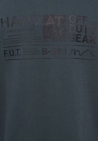 Mørk teal t-shirt med grafisk print, der indeholder tekst i varierende størrelser og stilarter, herunder "HALO" og "OFF DUTY GEAR", samt en lineær designdetalje.