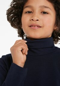 Marineblaues Rollkragenoberteil aus weichem Stoff, mit langen Ärmeln und umgeschlagenem Kragen. Die Hand des Modells befindet sich in der Nähe des Kragens.