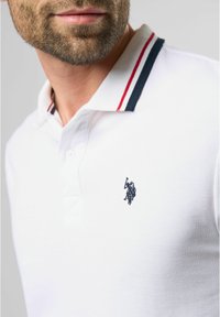 Hvid polo shirt med en struktureret overflade, prydet med et marineblåt hestelogo og en krave med røde og marineblå striber.
