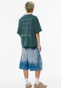 Green oversized t-shirt with a white heart pattern on the back, paired with light blue denim shorts and beige sneakers. White crew socks visible.