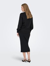 Moteris su ilgais blondiniais plaukais, vilkinti juodą megztinį, juodą ribbančią midi sijoną ir žemakulnius juodus slingback batus, stovinti šonu prieš pilką foną.