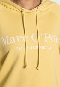 Hoodie in cotone giallo con il testo bianco "Marc O'Polo" e "EST. IN STOCKHOLM", dotato di cappuccio con cordini e polsini a costine.