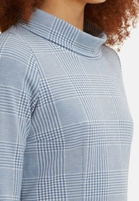 Camiseta de manga larga en azul claro con cuello drapeado. Presenta un patrón a cuadros con texturas y formas variadas, mostrada desde un lado.